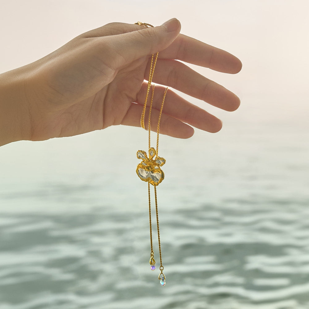Risis Oncidium Coco Drop Slider Necklace With Crystal Tailends Pg - A hand holds a delicate gold necklace with a translucent flower pendant and dangling chains ending in iridescent teardrop gems, exuding elegance and finesse.