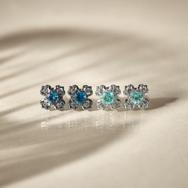 Risis Peranakan Kebaya Necklace Rh With Sky Blue Topaz 1 - Three pairs of floral earrings with silver petals and central gemstones in varying shades of blue sparkle against a soft surface.