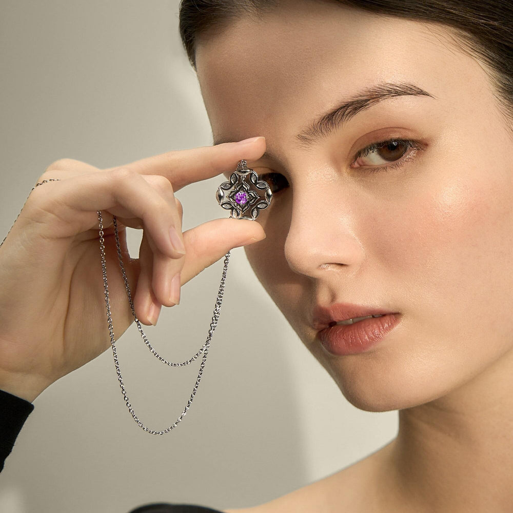 Risis Copy Of Peranakan Moments Darling Necklace With Amethyst - A silver, ornate pendant with a central purple gem is being held between fingers, suspended on a delicate chain near a woman's face.