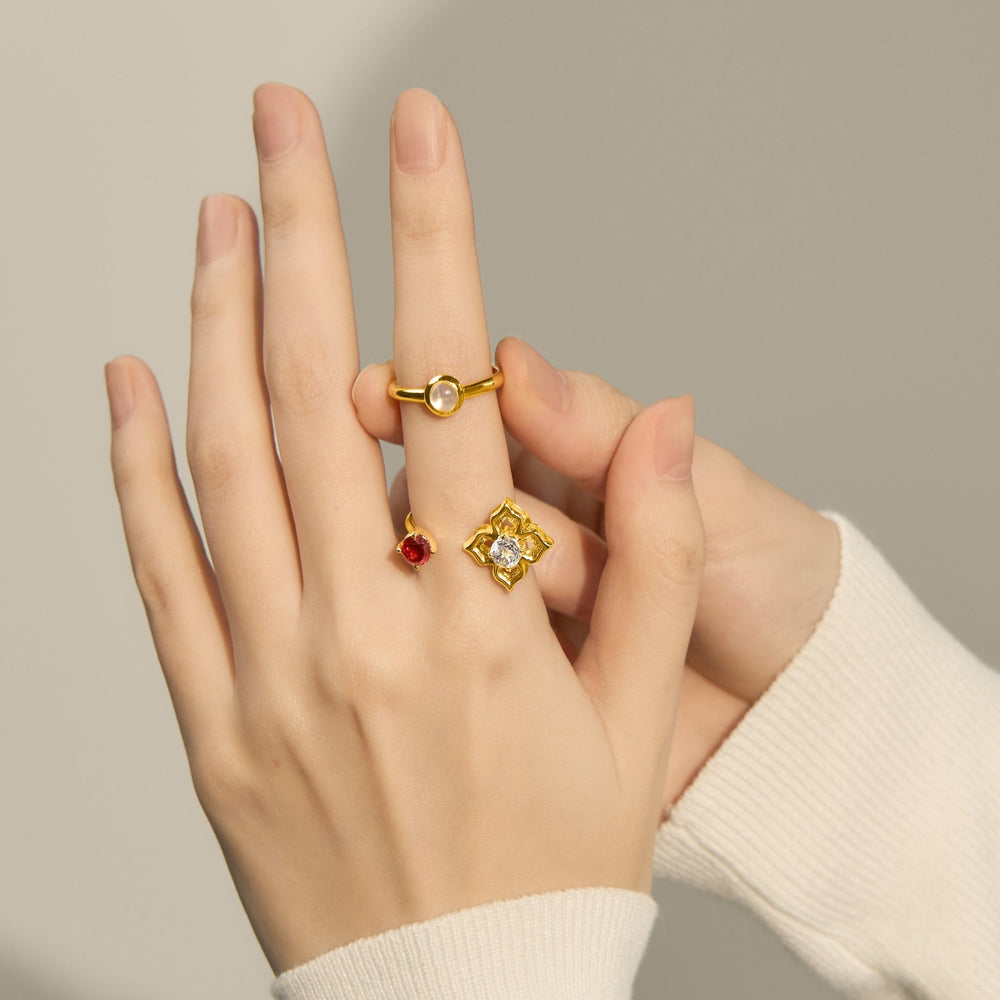 Risis Peranakan Moments Lace Ring with Red Garnet and White Topaz - Two gold rings adorn a person's fingers; one features a central red gemstone, while the other showcases a clear, faceted stone surrounded by intricate patterns.