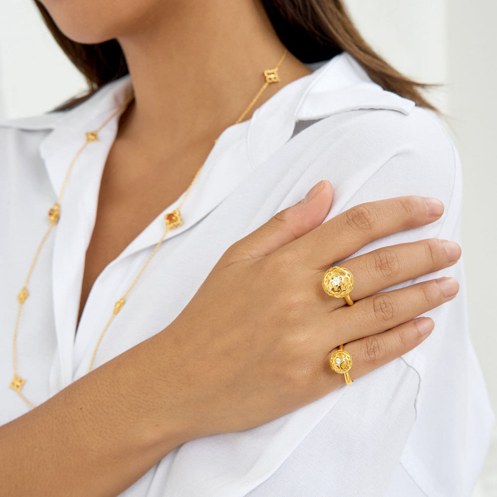 Risis Peranakan Spheres Large Ring with White Topaz (G) - A person is wearing gold rings with intricate designs and a delicate gold necklace embellishing a white shirt, conveying elegance and style.