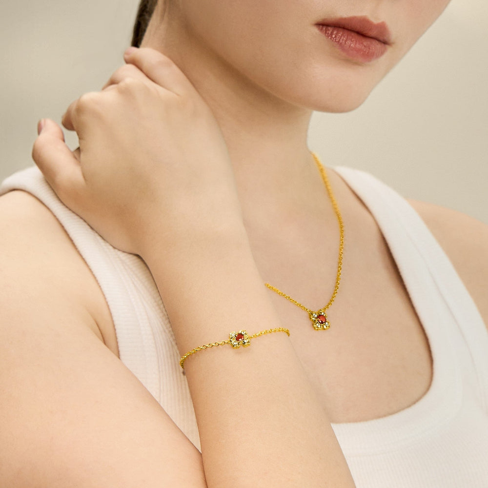 Risis Petite Peranakan Kebaya Necklace With Garnet - A delicate gold necklace drapes around a neck with a small, red gem pendant; a matching bracelet adorns the wrist, showcasing similar vibrant stones.