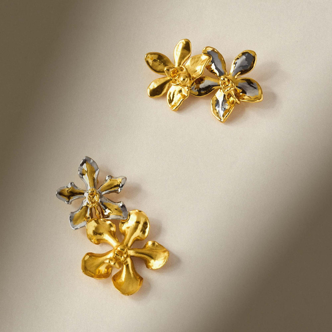 Risis Vanda And Kagawara Orchid Brooch - Three-dimensional, flower-shaped earrings with polished gold petals and contrasting, reflective silver embellishments rest elegantly against a muted surface.