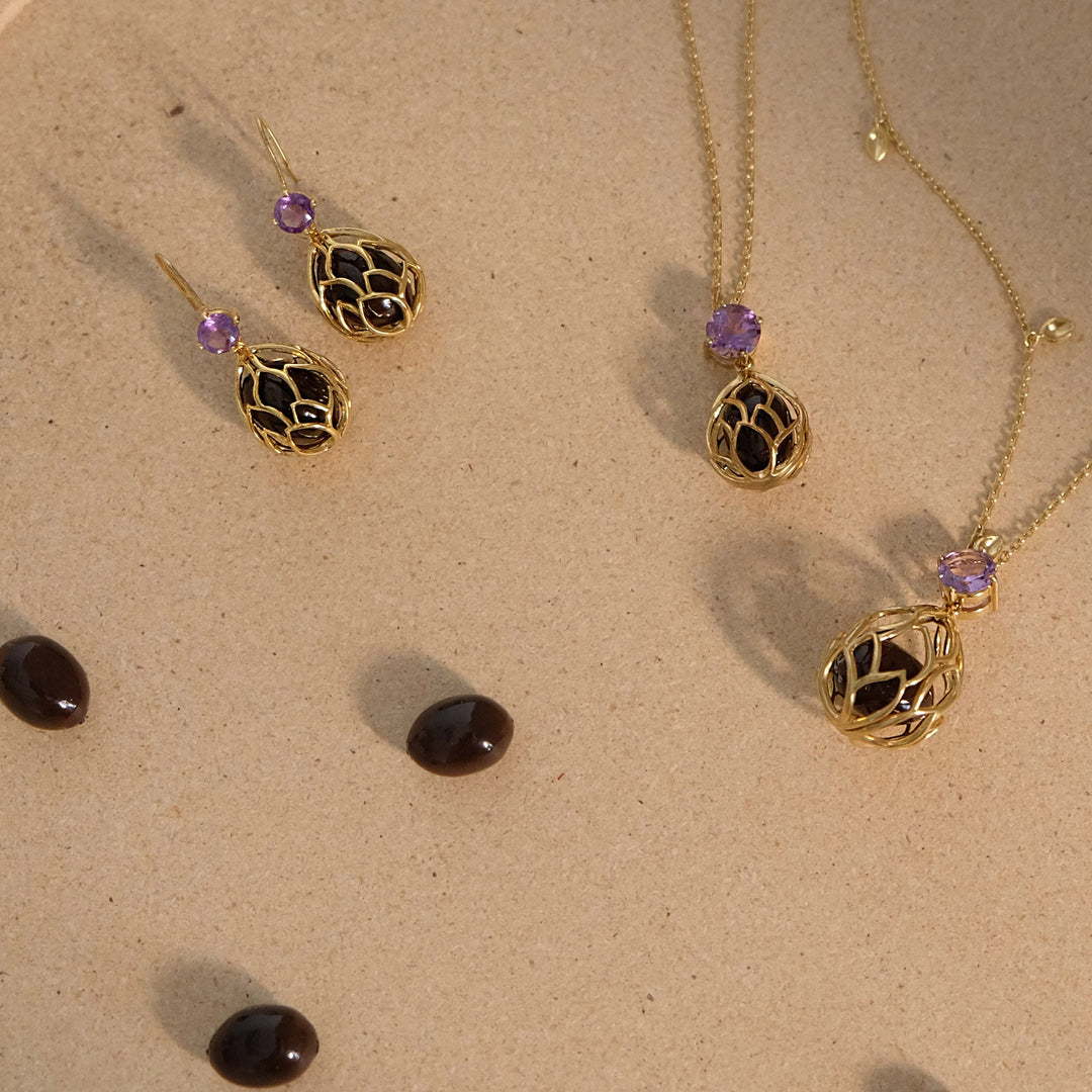 Risis Earrings Lotus Seeds - Gold jewelry with intricate cage-like pendants and earrings, each adorned with a small, round, purple gemstone, is elegantly displayed on a beige surface.