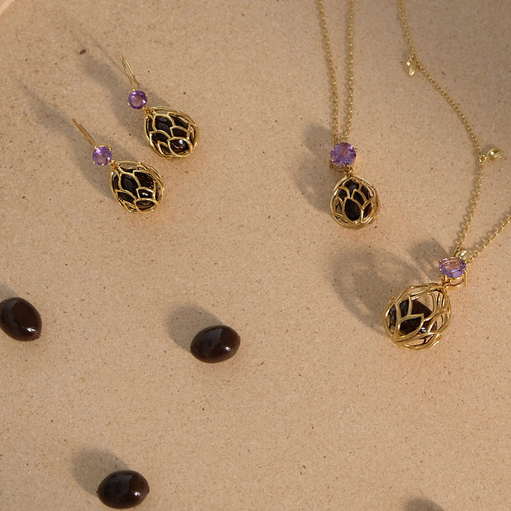 Risis Earrings Lotus Seeds - Gold jewelry with intricate cage-like pendants and earrings, each adorned with a small, round, purple gemstone, is elegantly displayed on a beige surface.