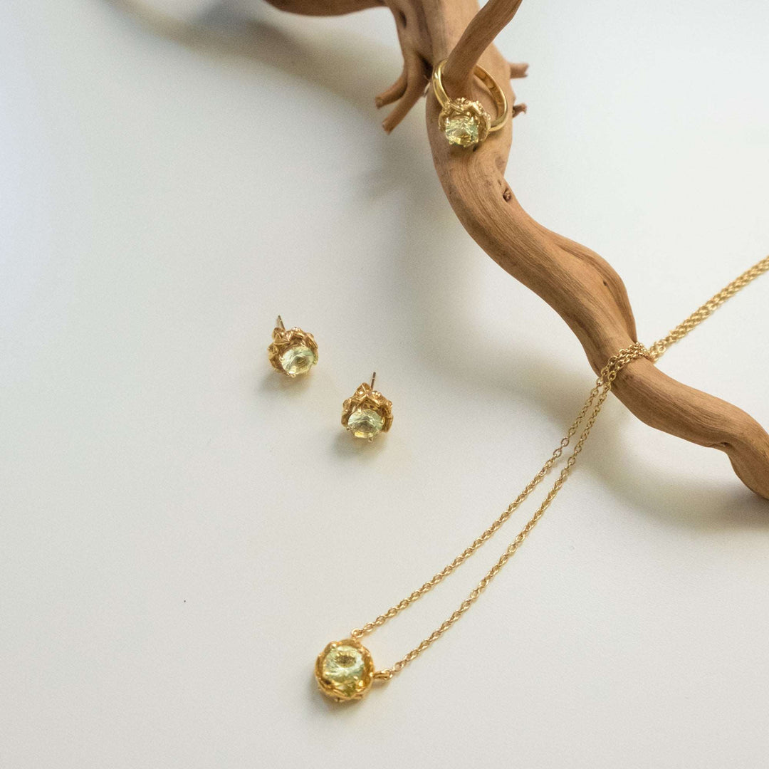 Risis Midori Earrings with Prasiolite - Golden jewelry set with an amber luster comprising a pendant necklace and matching earrings rests against a wooden form, exuding rustic elegance.