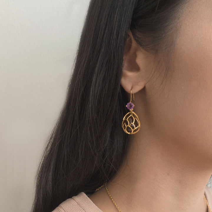 Risis Earrings Lotus Seeds - A gold earring with intricate looping designs and a small purple bead dangles from a woman's ear, complementing her sleek dark hair.