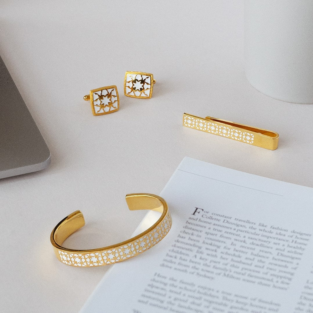 Risis Peranakan Orchid Tie Bar with White Enamel - Gold jewelry with intricate patterns, including cufflinks, a tie clip, and an open bangle, elegantly poised beside printed text.