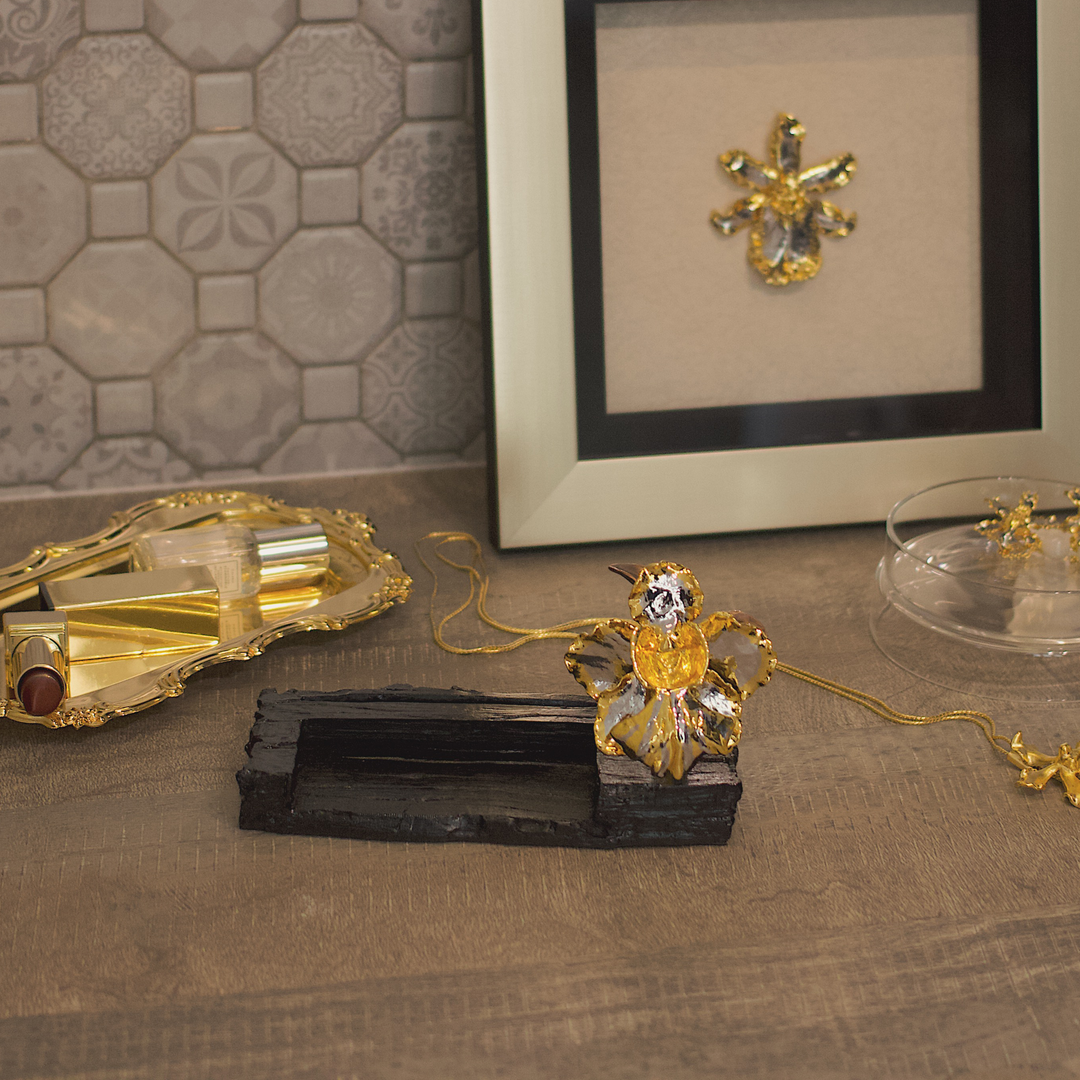 Risis Vanda Miss Joaquim Orchid Card And Pen Holder Charcoal Rhg - Golden jewelry with amber tones on a wooden display; a framed ornament is in the background.