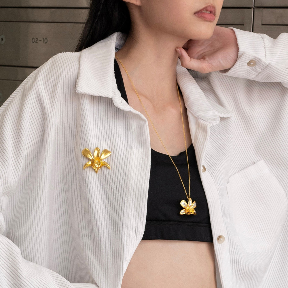 Risis Dendrobium Li Min Eng Orchid Brooch/Pendant (G) - A person is wearing a white shirt with a gold orchid-shaped brooch and a matching pendant necklace against a black top. The jewelry gleams with elegance.