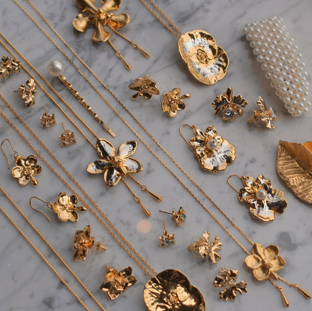 Risis Peranakan Earrings With White Topaz G - An assortment of gold-toned jewelry with floral and leaf designs, pearls, and a mix of polished and textured finishes, shimmering on a marble surface.