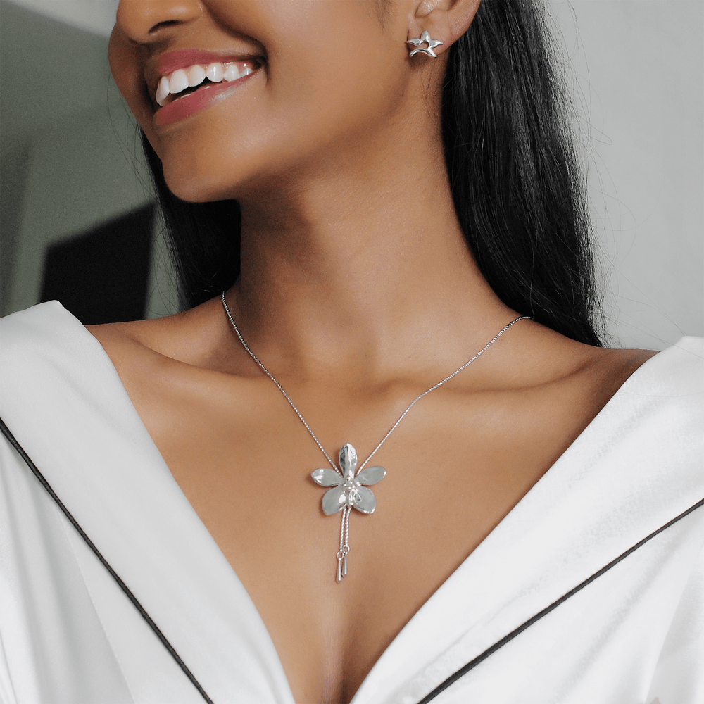 Risis Aeridovanda Vieng Ping Orchid Slider Necklace (P) - A silver necklace with a flower pendant, featuring shimmering petals and a drop charm, adorns a person's neck, complemented by matching star-shaped earrings.