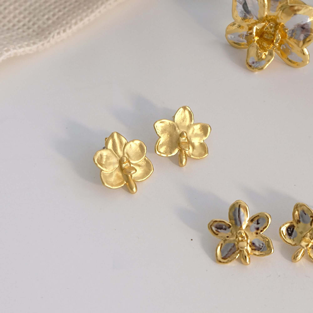 Risis Ascocenda Sagarik Gold Orchid Earrings (G) - The image shows shiny gold earrings shaped like four-petaled flowers, some with translucent gem petals and yellow centers, arranged asymmetrically on a flat surface.