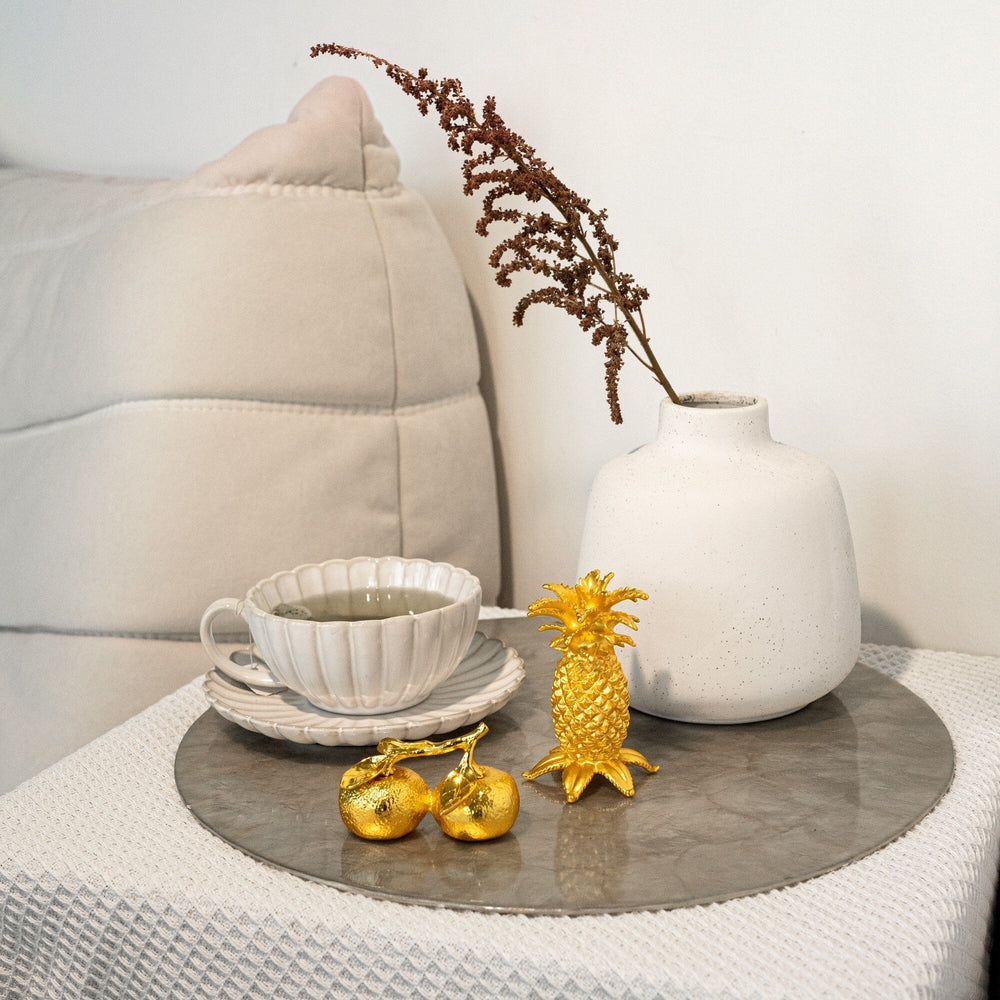 Risis Auspicious Golden Tangerines - A white ceramic vase with a brown plant sprig, beside sculptural golden fruit ornaments, atop a gray circular surface; nearby, white stacked teacups add a domestic touch.