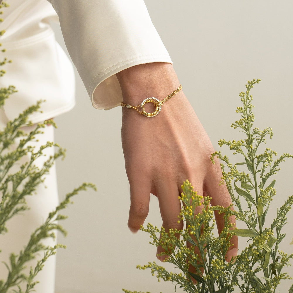 Risis Bamboo Twist Bracelet with White Sapphires - A golden bracelet with an intricate, hollow circular charm adorns a delicate wrist, complemented by slender fingers gracefully resting among soft green foliage.