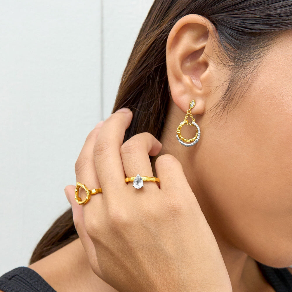 Risis Bamboo Twist Dangling Earrings with White Sapphires - A person is wearing gold jewelry: a dangling earring with encrusted gems and two rings, one with a clear teardrop gemstone, expressing elegance and style.