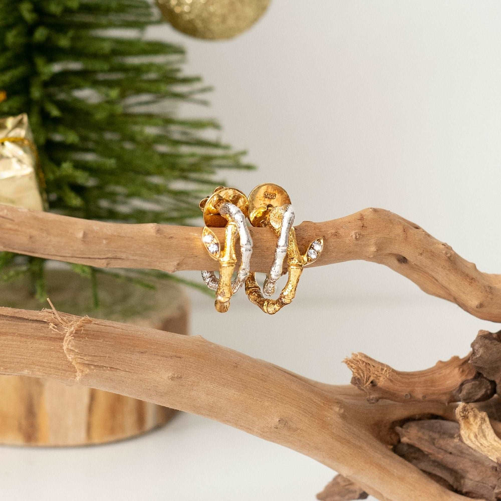Risis Bamboo Twist Pierced Earrings with White Sapphires - A pair of gold-tone earrings with a twisted design and encrusted rhinestones, displayed on a natural wood branch.