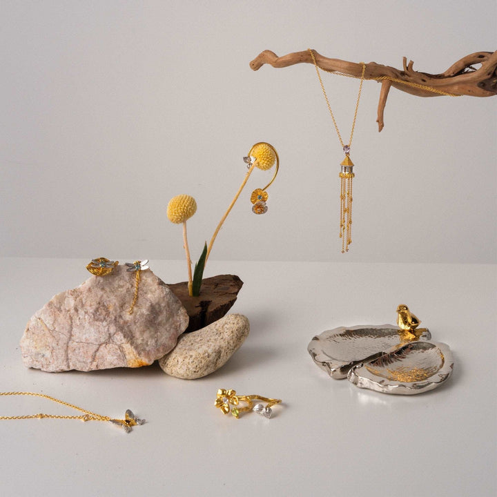 Risis Botanique Butterfly Necklace - Elegant jewelry with gold tones and gem embellishments is artistically arranged among rocks and a dried plant, displaying a blend of natural and crafted beauty.