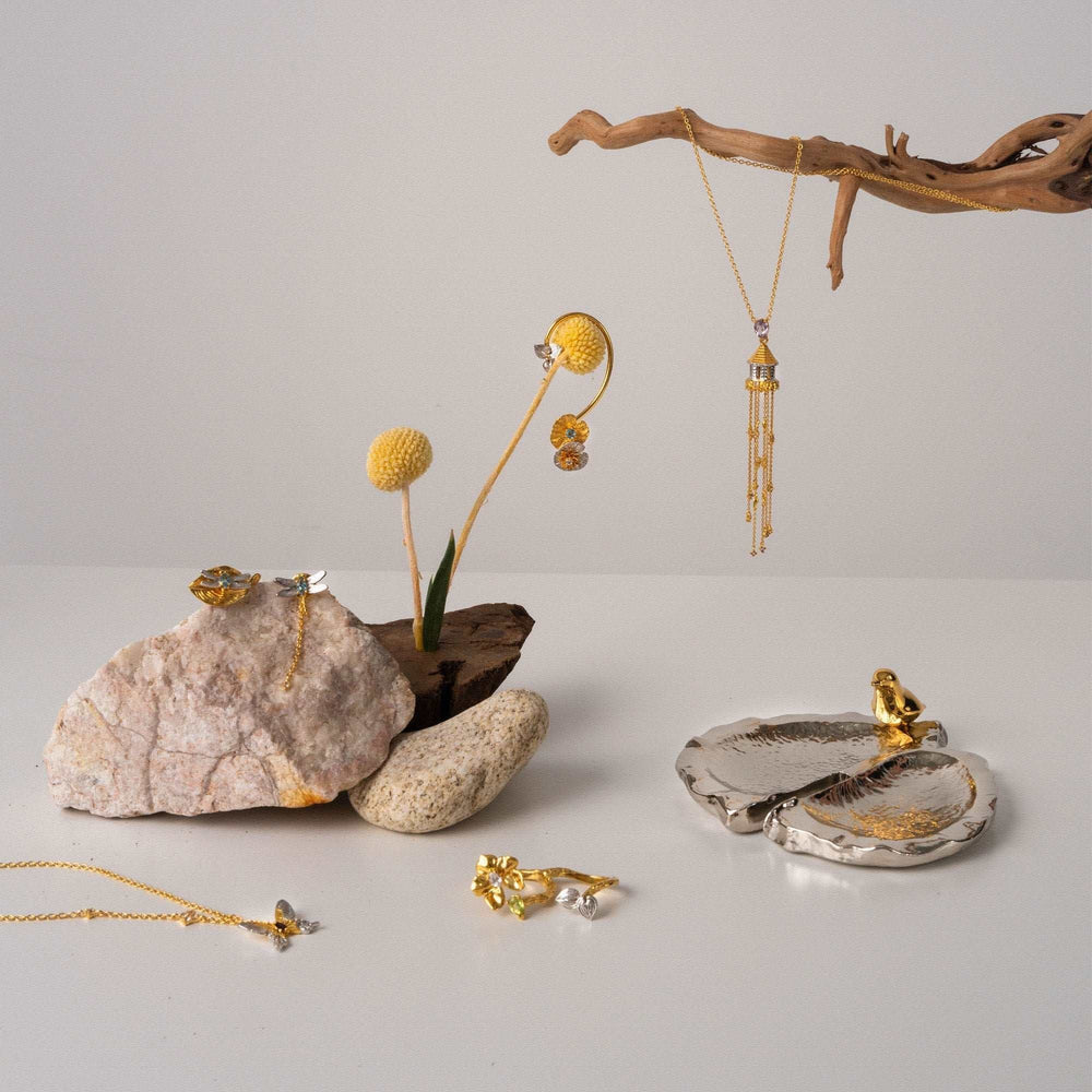 Risis Botanique Orchid Ring - Intricate golden jewelry with amber and crystal accents is artfully displayed among rocks and twigs, featuring earrings, necklaces, and rings with floral and geometric designs.