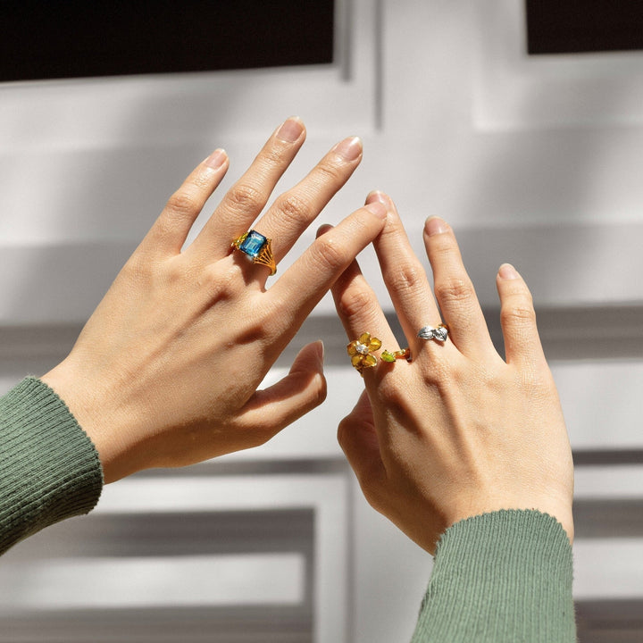 Risis Botanique Orchid Ring - Two hands adorned with four rings; featuring large gemstones in blue, yellow, and clear hues, set in reflective metallic bands, interacting with sunlight to accentuate their vibrant colors and sparkle.
