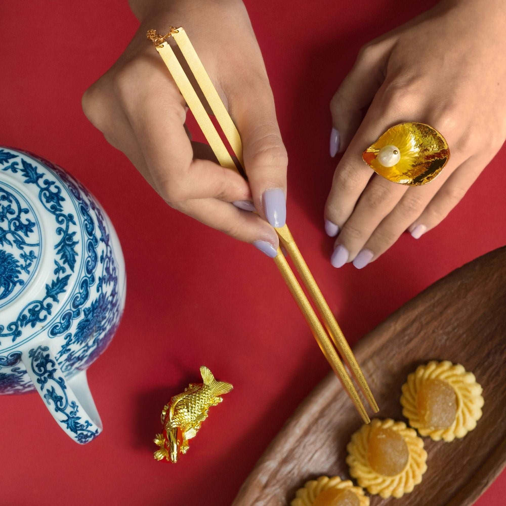 Risis Chopsticks with Carp Rest - A pair of hands with lavender nail polish holds elegant gold chopsticks, wears a large, amber-hued ring with a pearl inset, and is accompanied by a golden fish figurine.