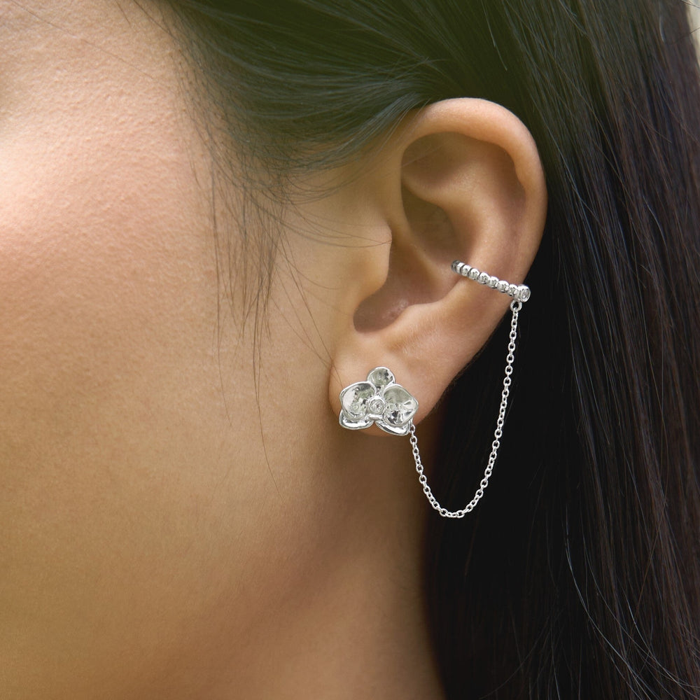 Risis Dainty Phalaenopsis Ear Cuff - A silver earring with a flower design and a chain connecting two parts shines against the ear of a person with black hair.