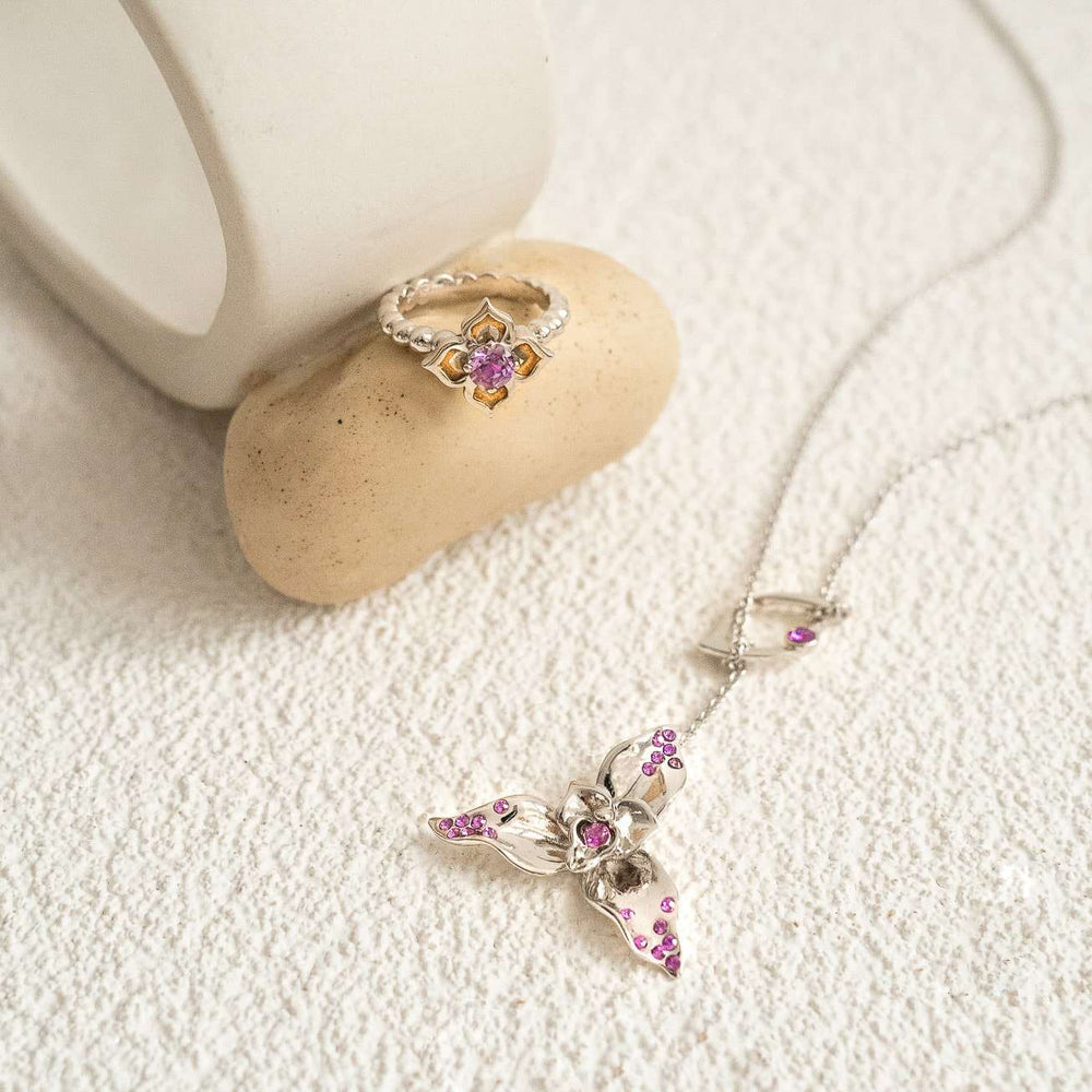 Risis Dazzle With Orchids Necklace - Lycaste Inspired Orchid with Amethyst (Feb) - A silver ring and pendant with pink gemstones rest on a pale surface, the ring on a small stone and the pendant with floral elements on the chain.