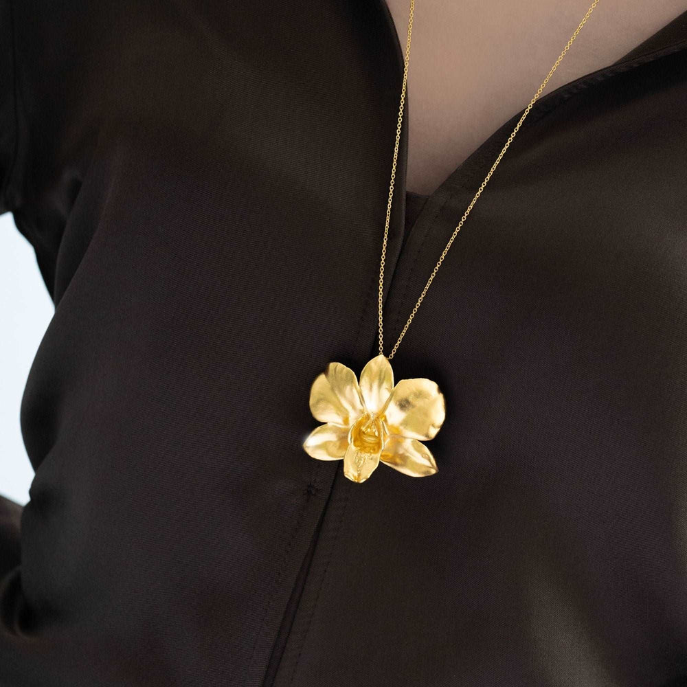 Risis Dendrobium Eleanor Chan Orchid Necklace (G) - A golden floral pendant hangs on a thin chain, set against a dark blouse. The flower has multiple petals, creating an elegant, sculpted appearance.