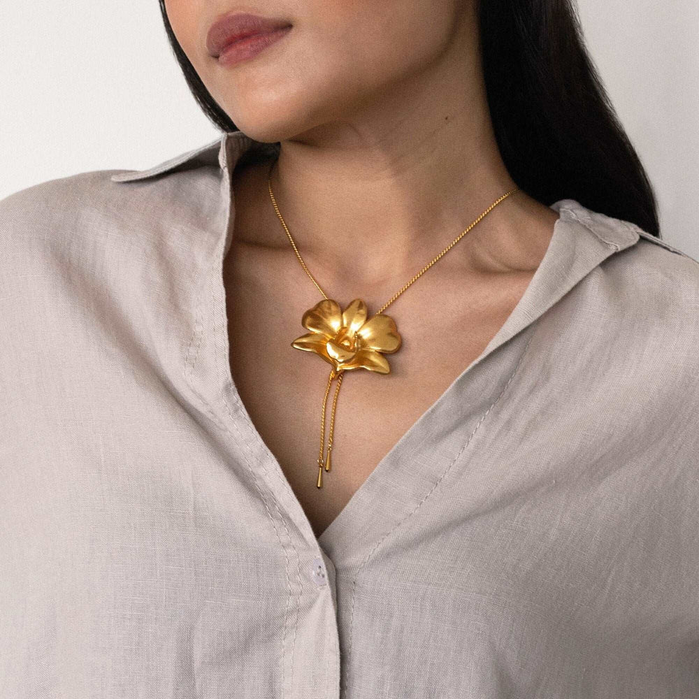 Risis Dendrobium Tay Swee Keng Orchid Slider Necklace (G) - A shiny gold necklace with a large, sculpted flower pendant rests on a person's collarbone, accentuating a gray blouse. The necklace exudes elegance and simplicity.