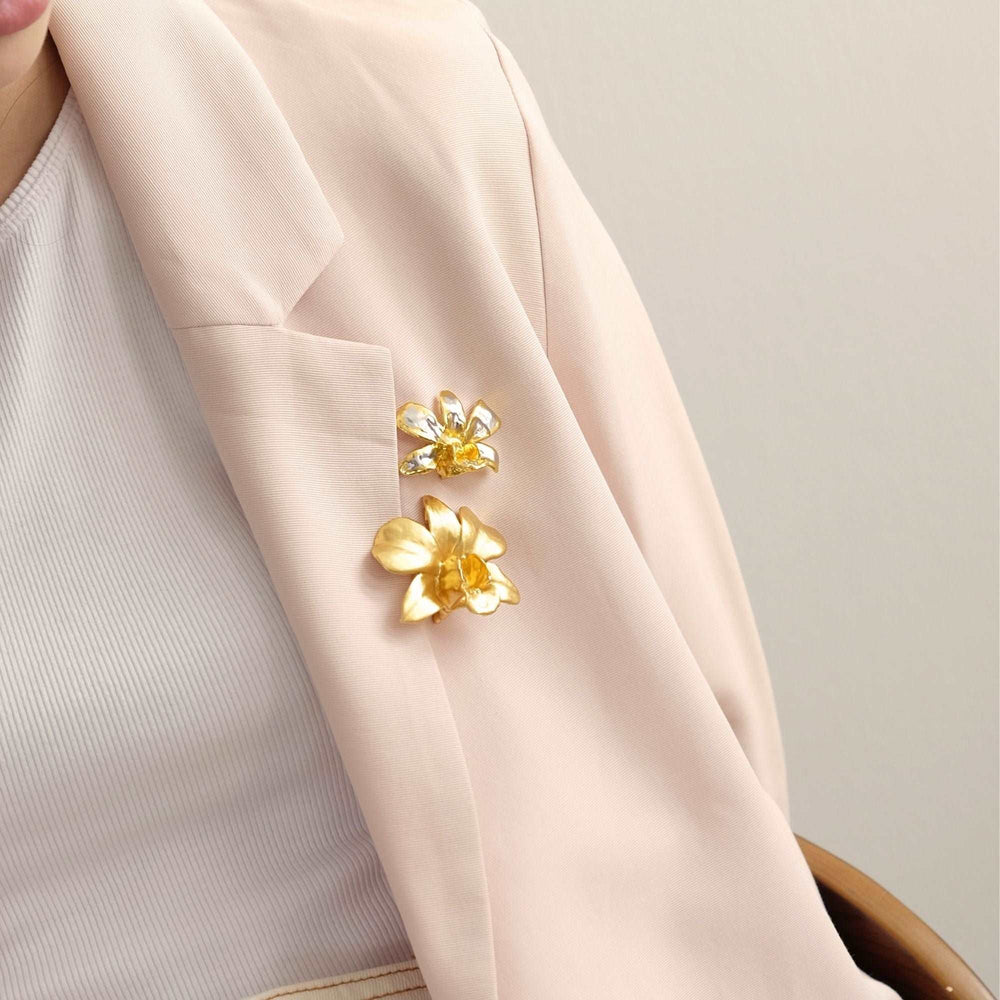 Risis Dendrobium Thong Chai Orchid Brooch/Pendant (PG) - A pair of golden floral brooches adorn the lapel of a pale pink blazer, each featuring central gemstones that add a touch of sparkle to their elegant design.