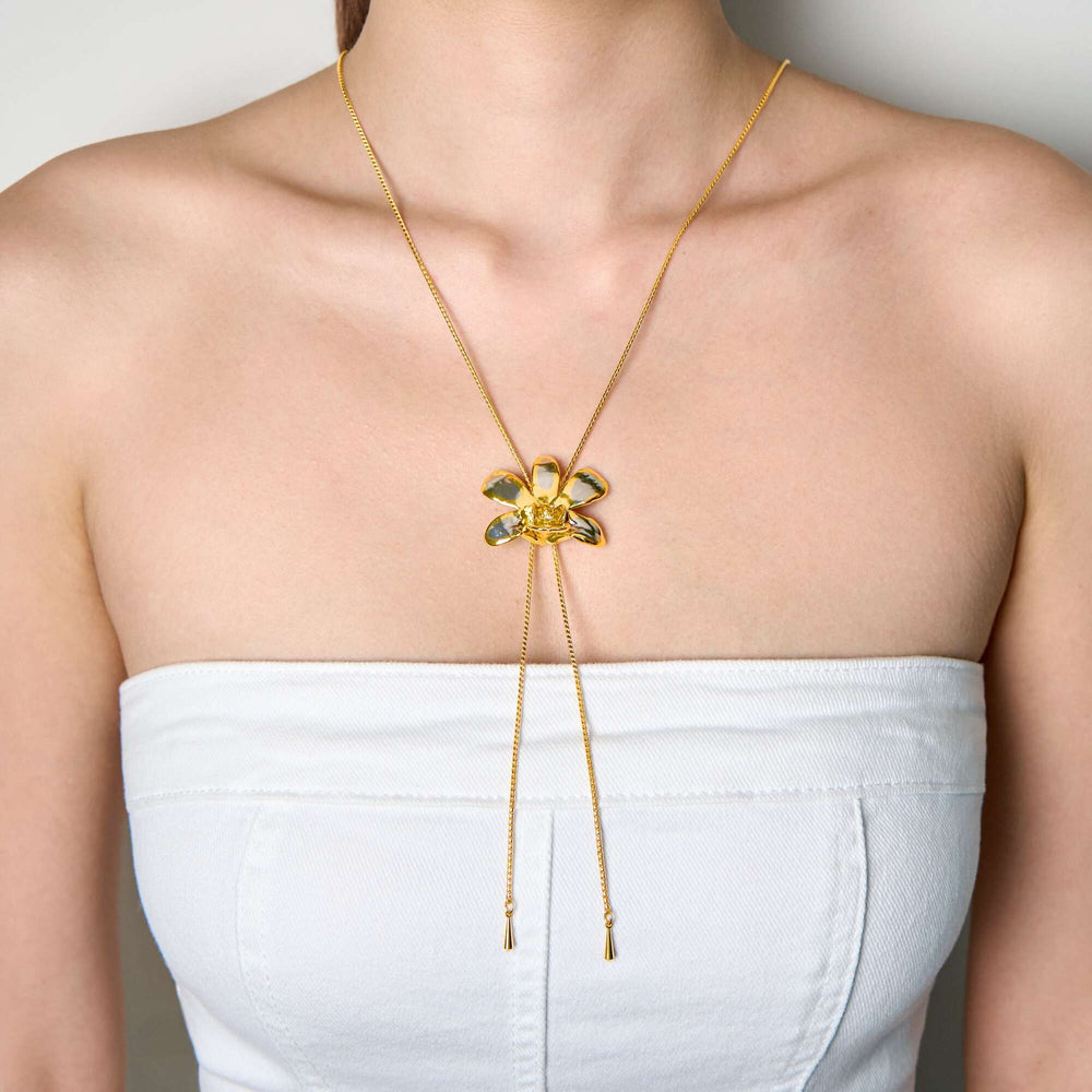 Risis Dendrobium Thong Chai Orchid Slider Necklace (PG) - A golden necklace with a delicate bow pendant adorned with translucent green gemstones hangs centered on a person's upper chest area.