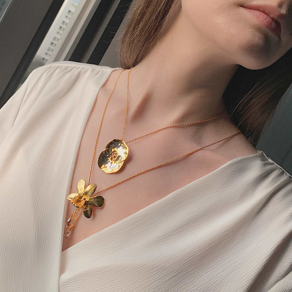 Risis Dendrobium Thong Chai Orchid Slider Necklace with Crystal Tailends (G) - A person is wearing two gold necklaces; one with a round, textured pendant and another with a floral motif and delicate gems.