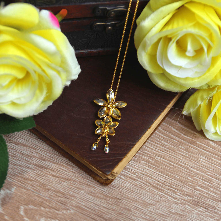 Risis Ascocenda Sagarik & Aeridovanda Vieng Ping Orchid Slider Necklace with Crystal Tailends (PG) - A golden cross pendant with a floral design and embedded clear gemstones hangs delicately against a dark book, flanked by vibrant yellow fabric flowers.
