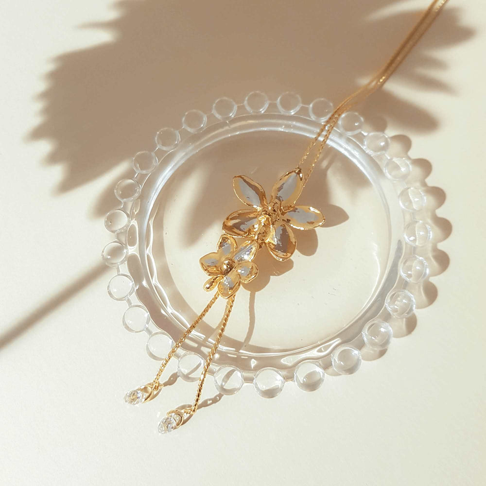 Risis Ascocenda Sagarik & Aeridovanda Vieng Ping Orchid Slider Necklace with Crystal Tailends (PG) - A golden necklace with a flower pendant and crystal embellishments rests elegantly on a clear, beaded glass coaster in bright natural light.