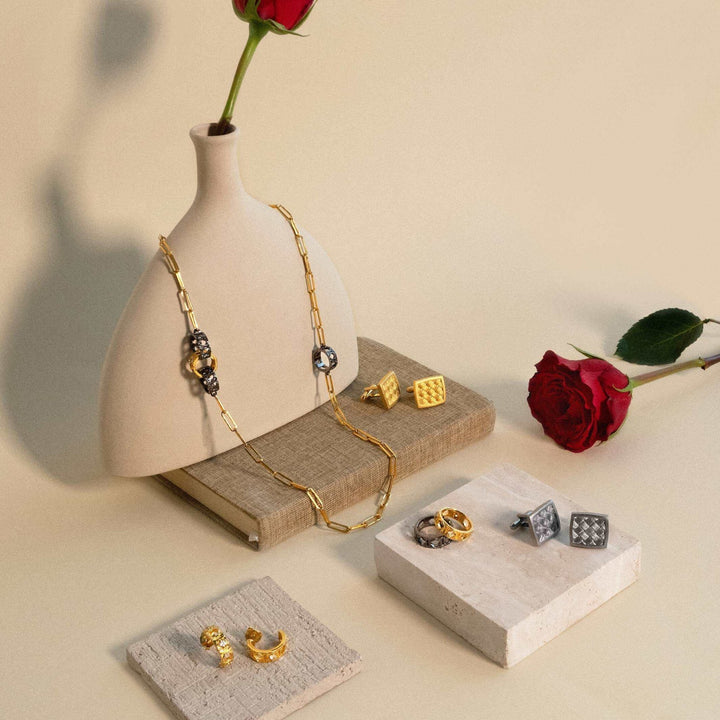 Risis Entwined Rattan Ring (BRH) - A gold chain necklace with ornate beads rests on a mannequin bust, accompanied by elegant rings and cufflinks displayed on stone tiles, alongside a single red rose.