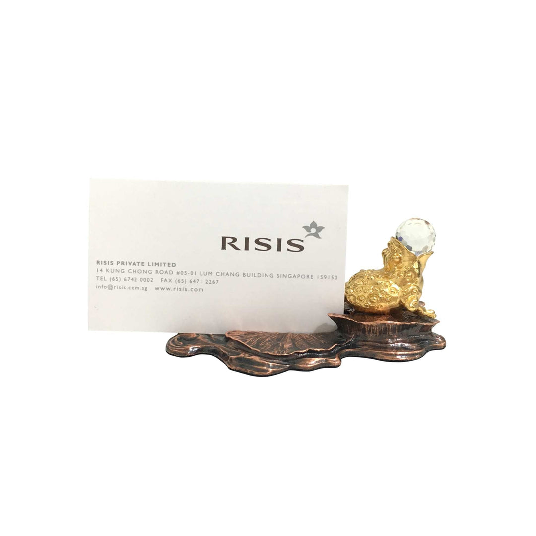 Risis Fortune Toad w Crystal on Lotus Leaf Name Card Holder - A golden sculpture of a frog with a clear orb on its back, resting on a copper-toned leaf-shaped base, reflecting craftsmanship and luxury.