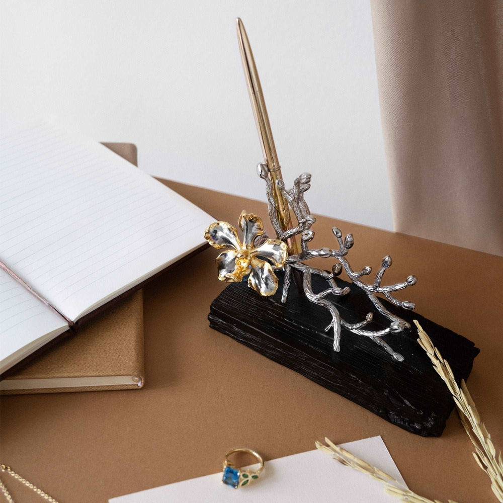 Risis Garden Odyssey Groves Pen and Name Card Holder - An elegant pen holder, crafted to resemble a silver branch with golden-bloomed flowers, rests atop a dark, textured base beside a ring with blue gem accents.