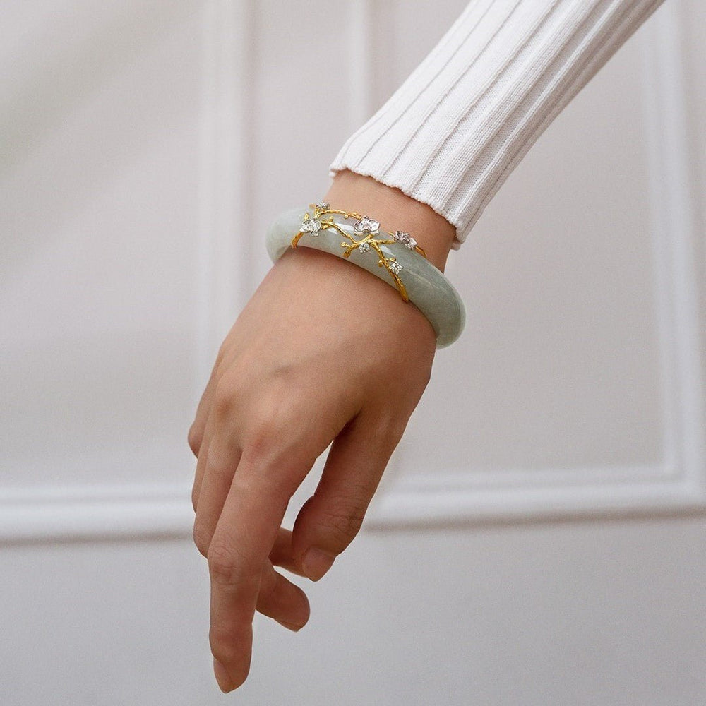 Risis Jadeite Bloom IV - A delicate silver bracelet features intricate branches with tiny, glossy gold and silver flowers, accented by sparkling gemstones, displayed against a soft green backdrop.