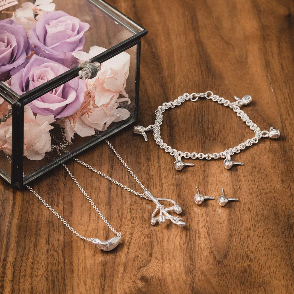 Risis Joyous Spring Earrings - A collection of silver jewelry, including a charm bracelet, pendant necklace, and earrings, is elegantly displayed on a wooden surface beside a box of pastel roses.