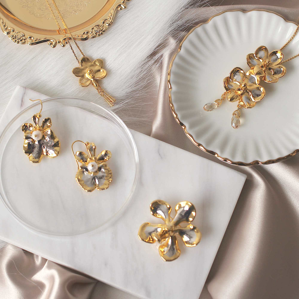 Risis Kagawara Firebird Orchid Brooch/Pendant (PG) - Golden floral jewelry with pearl accents lies gracefully, presenting earrings, a pendant, and a ring with an elegant aesthetic on a silky surface.
