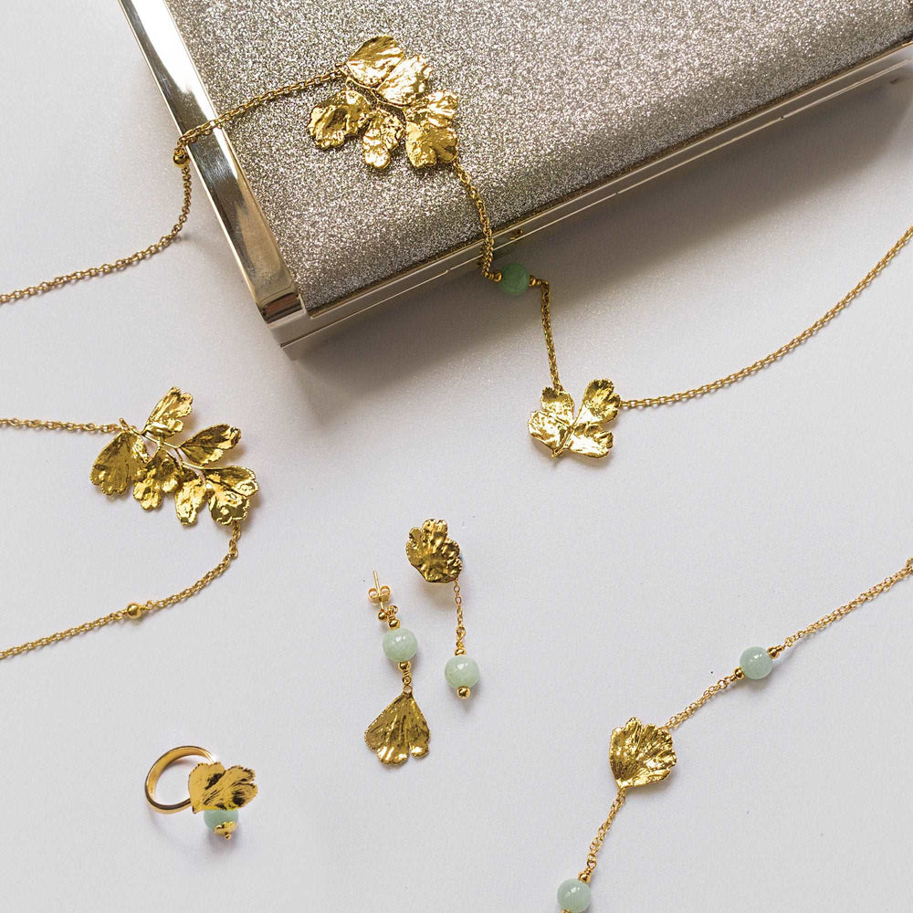 Risis Maiden Jade Earrings - Gold-toned jewelry with leaf motifs lays on a surface, including a necklace with small green beads, matching earrings, and a ring, all arranged near a glittery clutch.