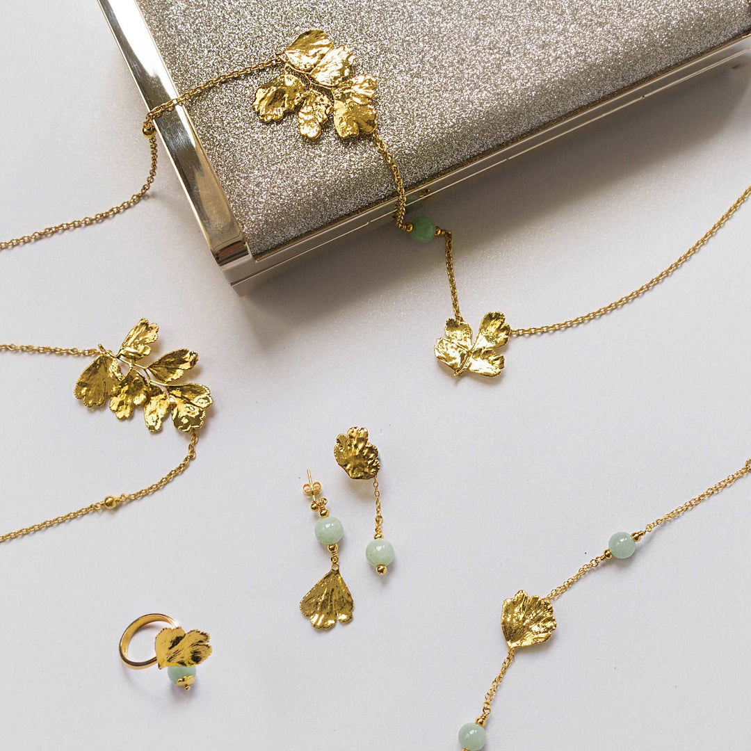 Risis Maiden Jade Earrings - Gold-toned jewelry with leaf motifs lays on a surface, including a necklace with small green beads, matching earrings, and a ring, all arranged near a glittery clutch.