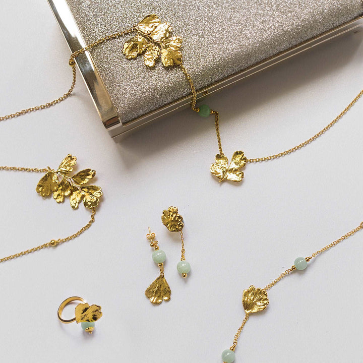 Risis Maiden Jade Earrings - Gold-toned jewelry with leaf motifs lays on a surface, including a necklace with small green beads, matching earrings, and a ring, all arranged near a glittery clutch.