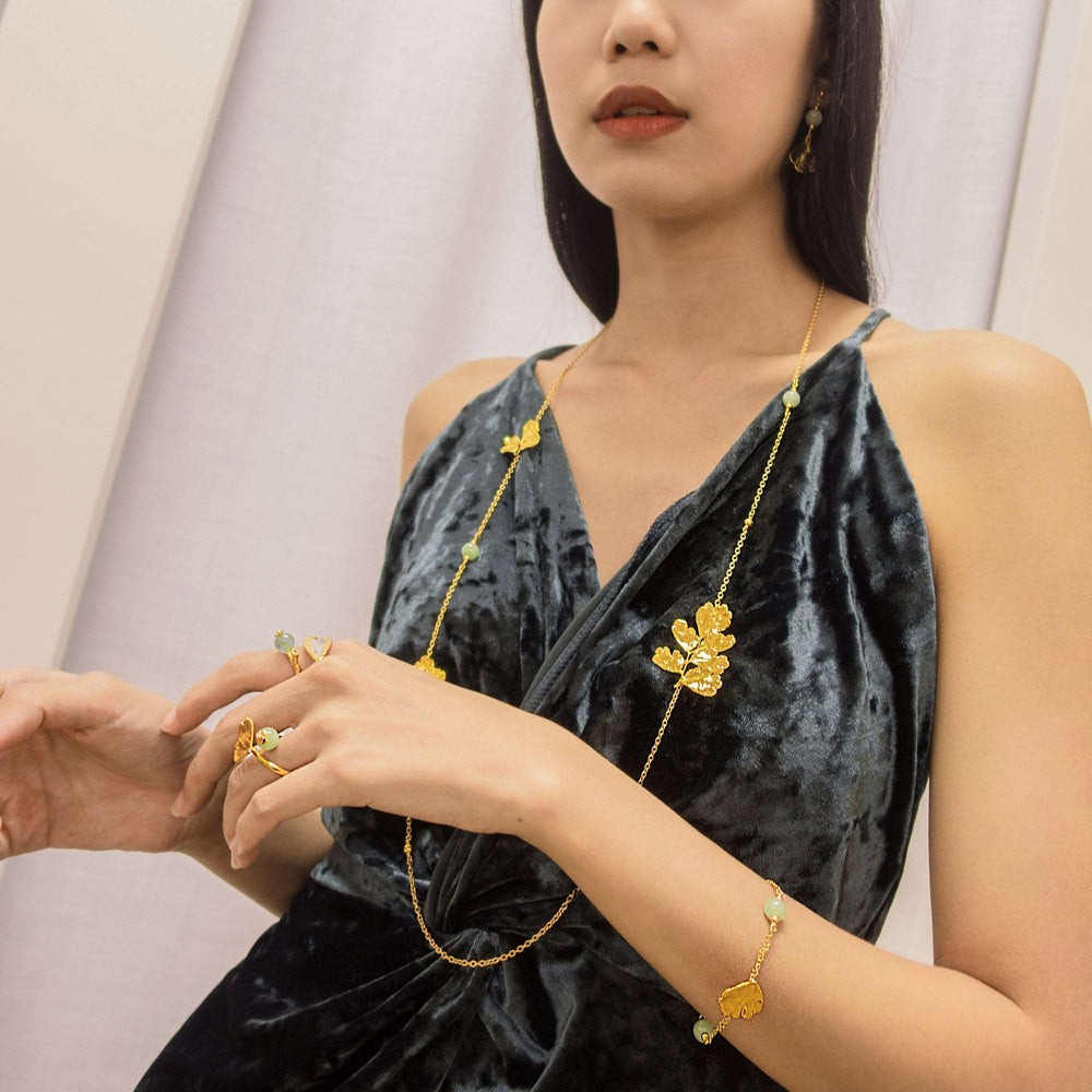Risis Maiden Jade Long Necklace - A person is adorned with golden jewelry featuring floral designs: delicate earrings, rings, a bracelet, and a necklace against a velvet garment.