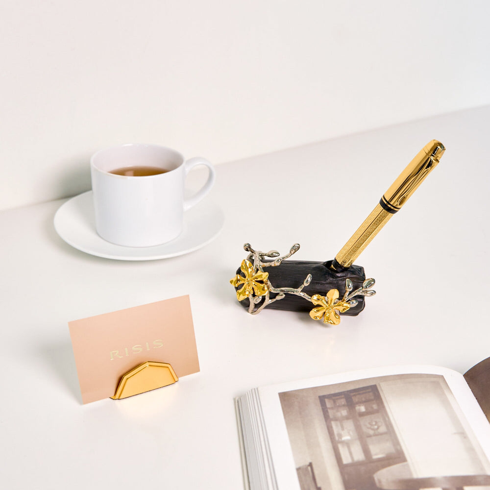 Risis Merlion Scale Name Card Holder - A gilded pen rests in an ornate holder with silver and gold flower embellishments, next to an open book, a white teacup on a saucer, and a peach-colored card.