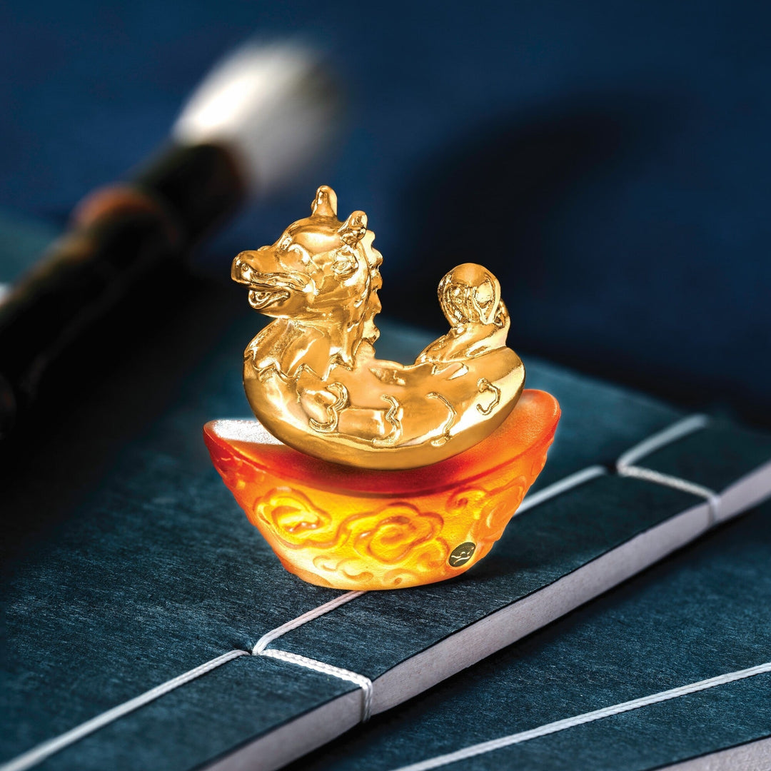 Risis Mini Celestial – Dragon on Amber Liu Li - A golden dragon and phoenix sculpture sits atop a translucent, orange, cloud-patterned base. Exquisite detailing highlights mythical elegance against a soft-focus backdrop.