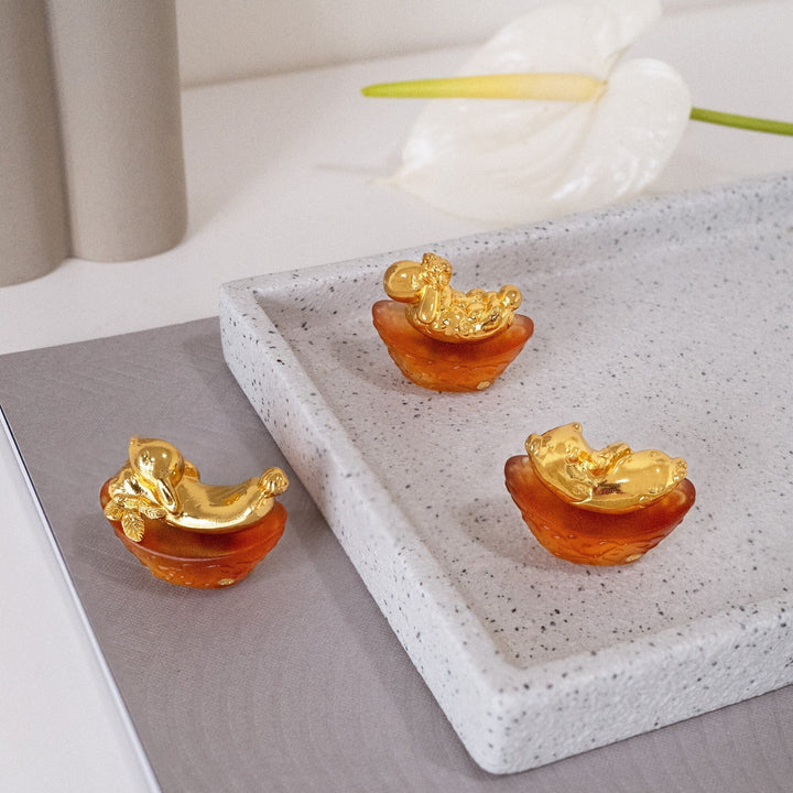 Risis Mini Celestial – Pig on Amber Liu Li - Three golden figurines resembling mythological creatures, with intricate details, are perched within translucent amber bowls on a textured gray surface.