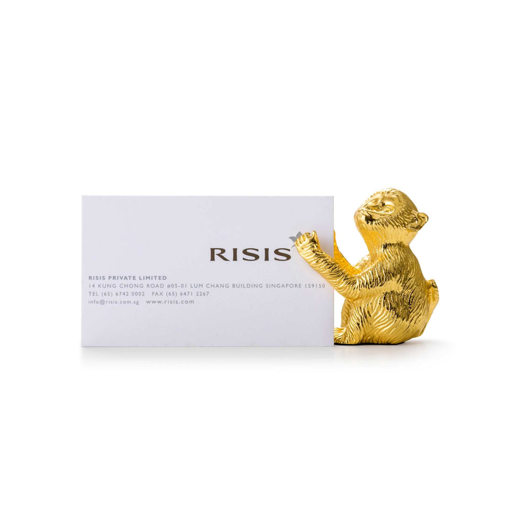 Risis Monkey - Influence Name Card Holder - A gold-tone figurine of a seated meerkat holds a business card prominently. The creature displays intricate textural details, imparting a sense of whimsy and elegance.