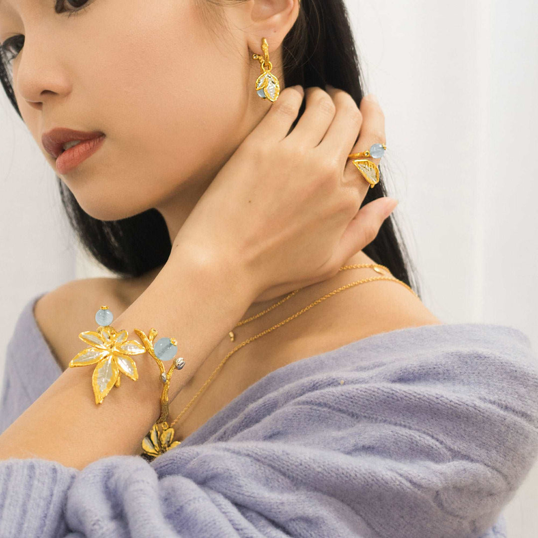 Risis Morning Glory Leaf Ring with Aquamarine and Topaz - A person is adorned with gold jewelry featuring leaf motifs and light blue gemstones—a bracelet, ring, and earrings, radiating elegance and harmony with a touch of nature's inspiration.