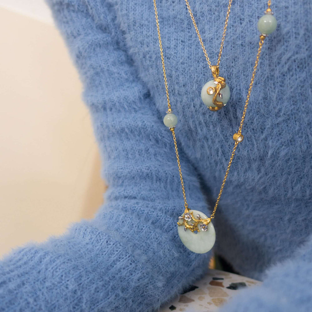 Risis Nature's Jade Long Necklace with White Topaz (L) - A layered gold necklace with pale green beads and a crescent moon pendant adorned with delicate flowers drapes over a soft blue sweater.